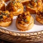 Toasted crostini topped with a caramelized apple mixture and creamy Brie, garnished with herbs on a white plate.