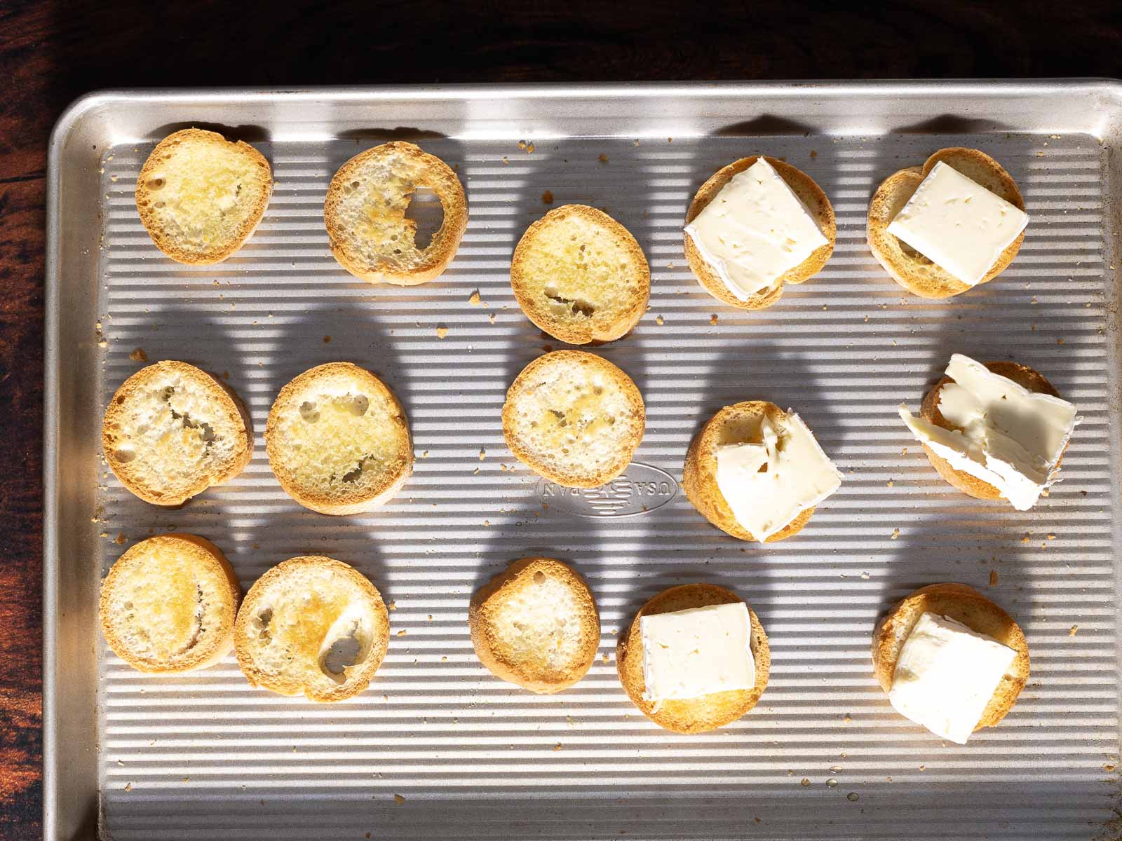 Slices of bread on a baking sheet, some with melted butter and others topped with creamy Brie cheese for a delightful crostini experience.