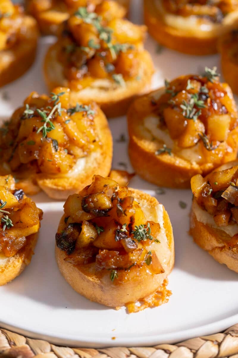 Close-up of crostini topped with caramelized apples, creamy brie, and herbs on a white plate.