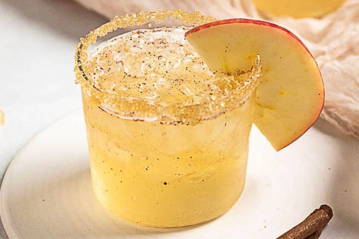A glass of yellow cocktail with ice, a sugared rim, and an apple slice garnish sits on a white plate-perfect for those who love to raise a glass to delicious Fall Cocktails.