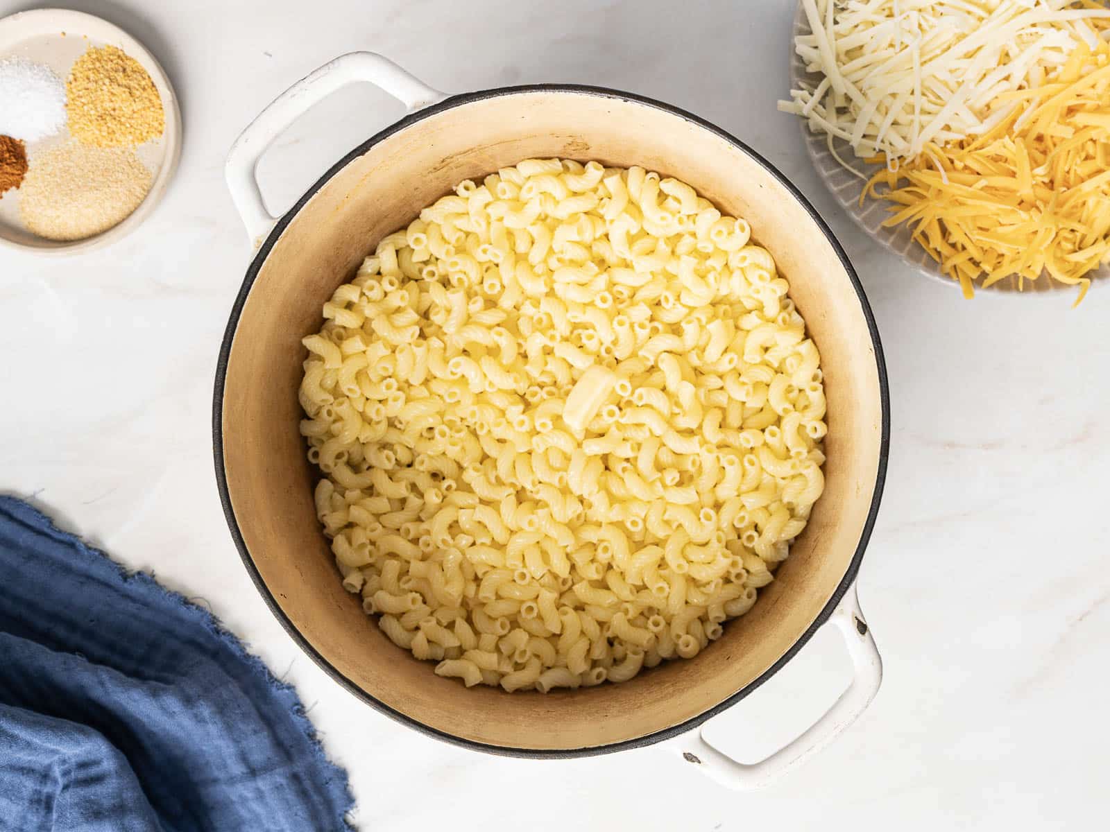 A pot of cooked elbow macaroni on a white surface, surrounded by bowls of shredded cheese and spices. A blue cloth is nearby.