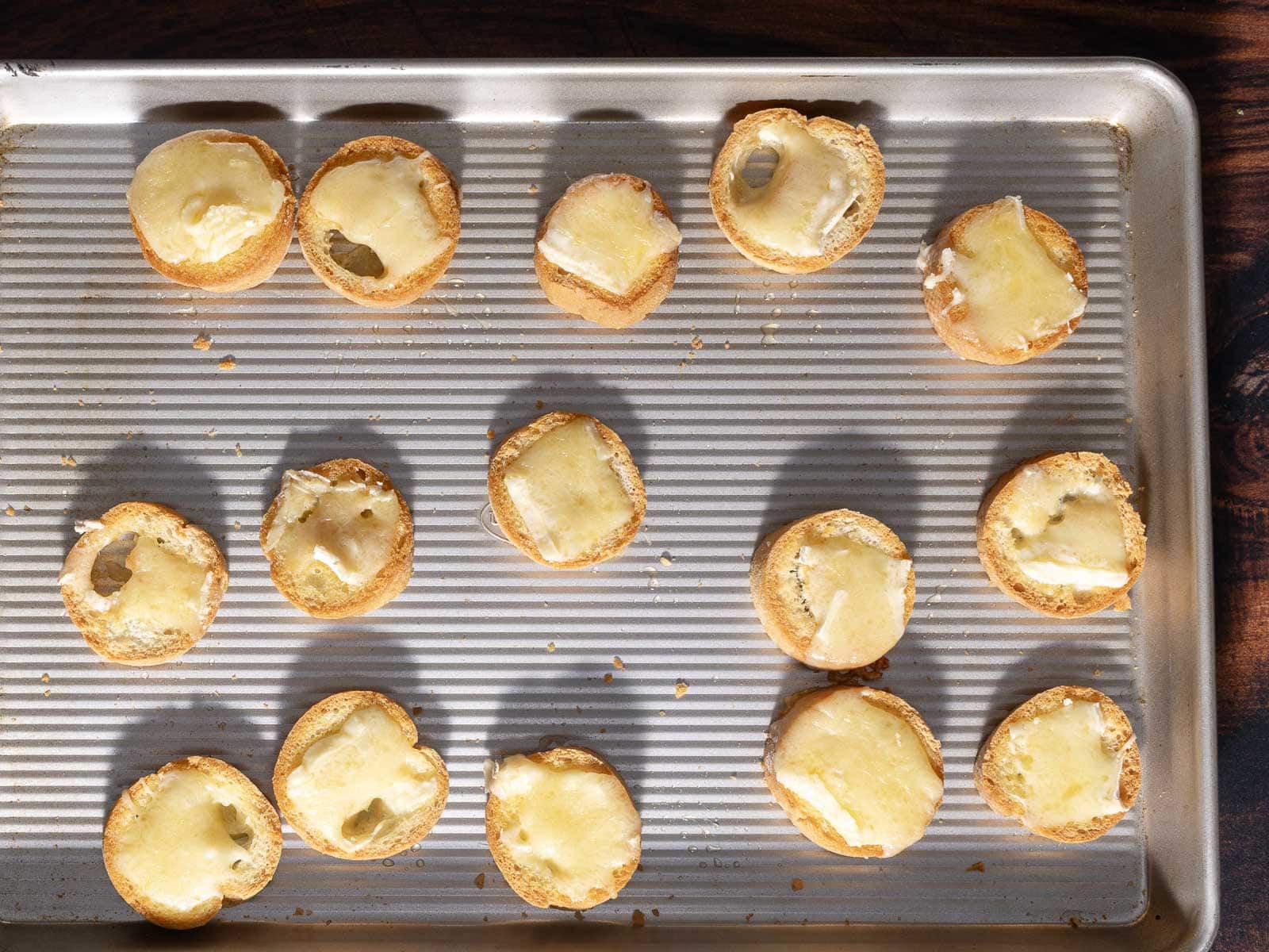 A baking tray lined with crostini topped with melted Brie cheese, each slice arranged neatly in rows.
