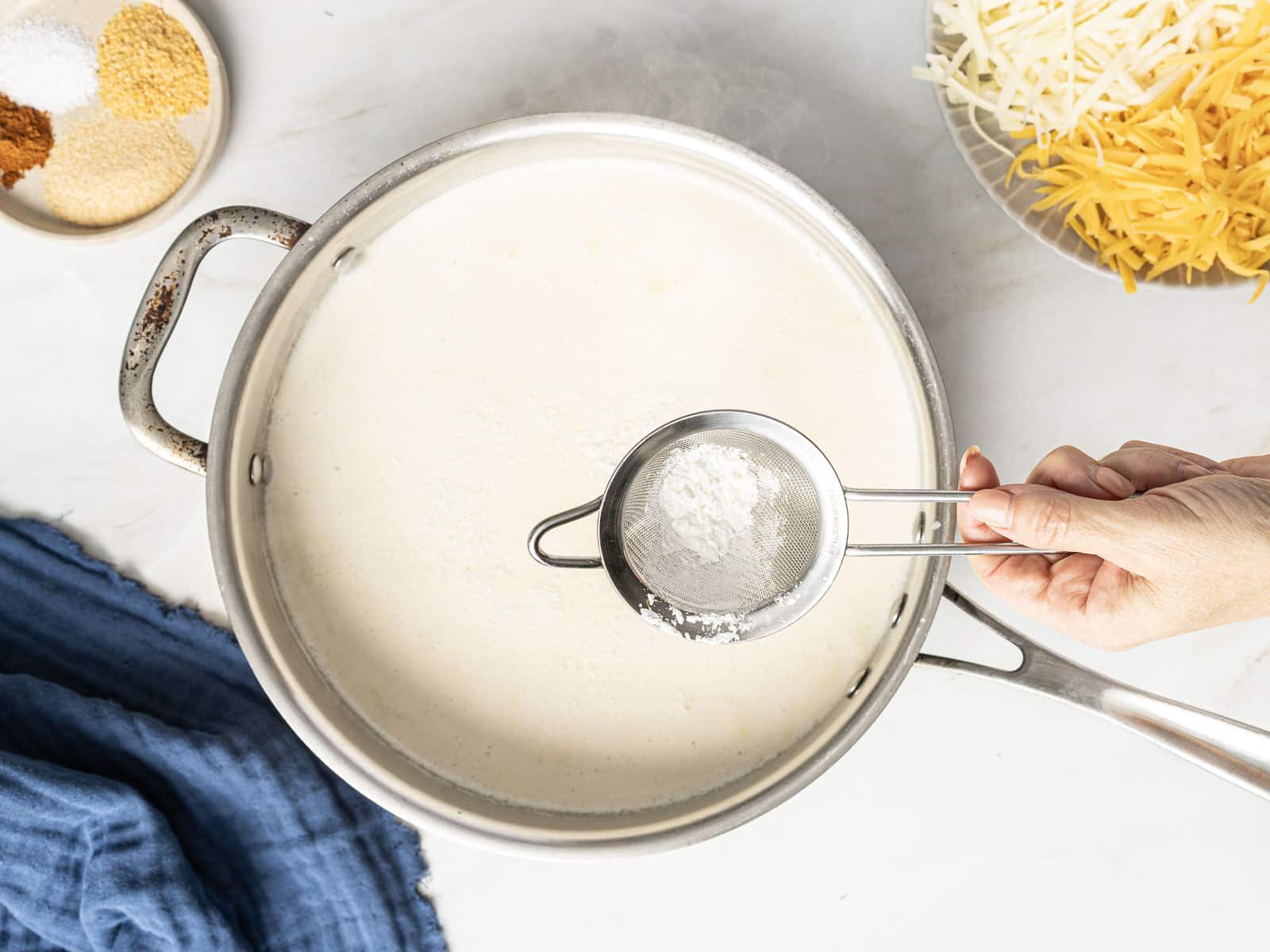 A hand holds a small sieve filled with flour over a pot of white sauce, surrounded by cheese and spices on a countertop.
