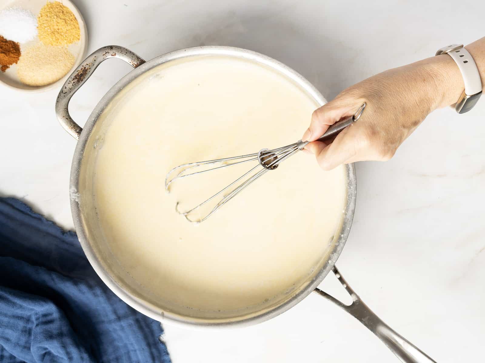A hand whisking a creamy sauce in a saucepan on a marble surface. A bowl with spices and a blue cloth are visible nearby.
