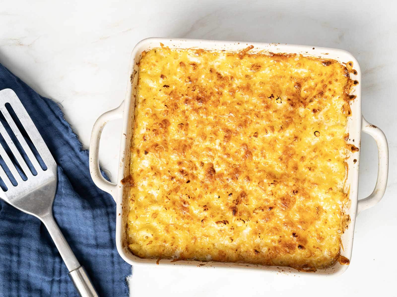 Square dish of golden-brown baked mac and cheese on a marble surface, next to a blue cloth and metal spatula.