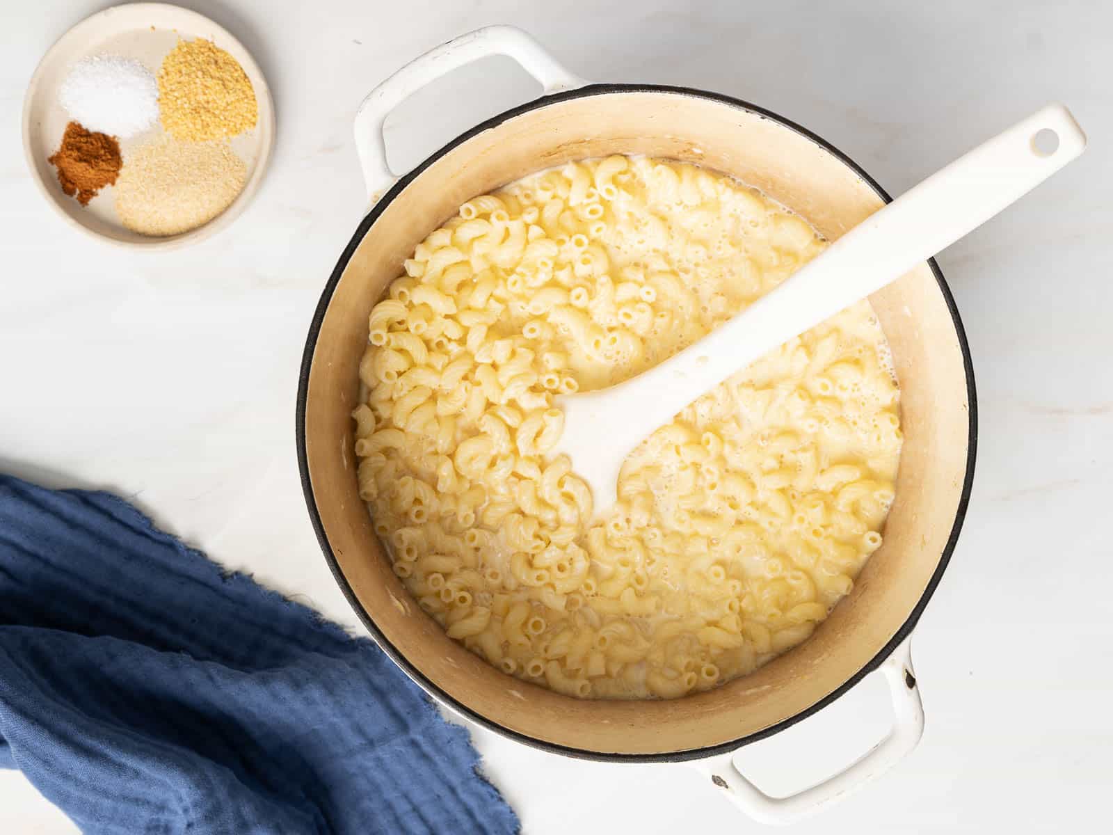Pot of cooked macaroni with a white spoon, next to a small dish with various spices, on a marble surface.