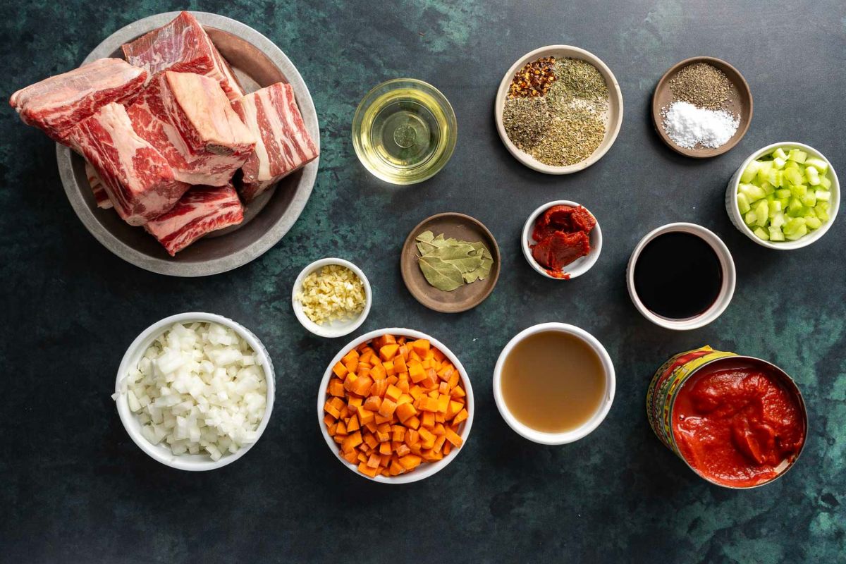Ingredients for a hearty Braised Beef Ragu featuring succulent beef ribs, chopped onions, carrots, celery, canned tomatoes, spices, broth, soy sauce, garlic, tomato paste, oil, and bay leaves elegantly arranged on a dark surface.