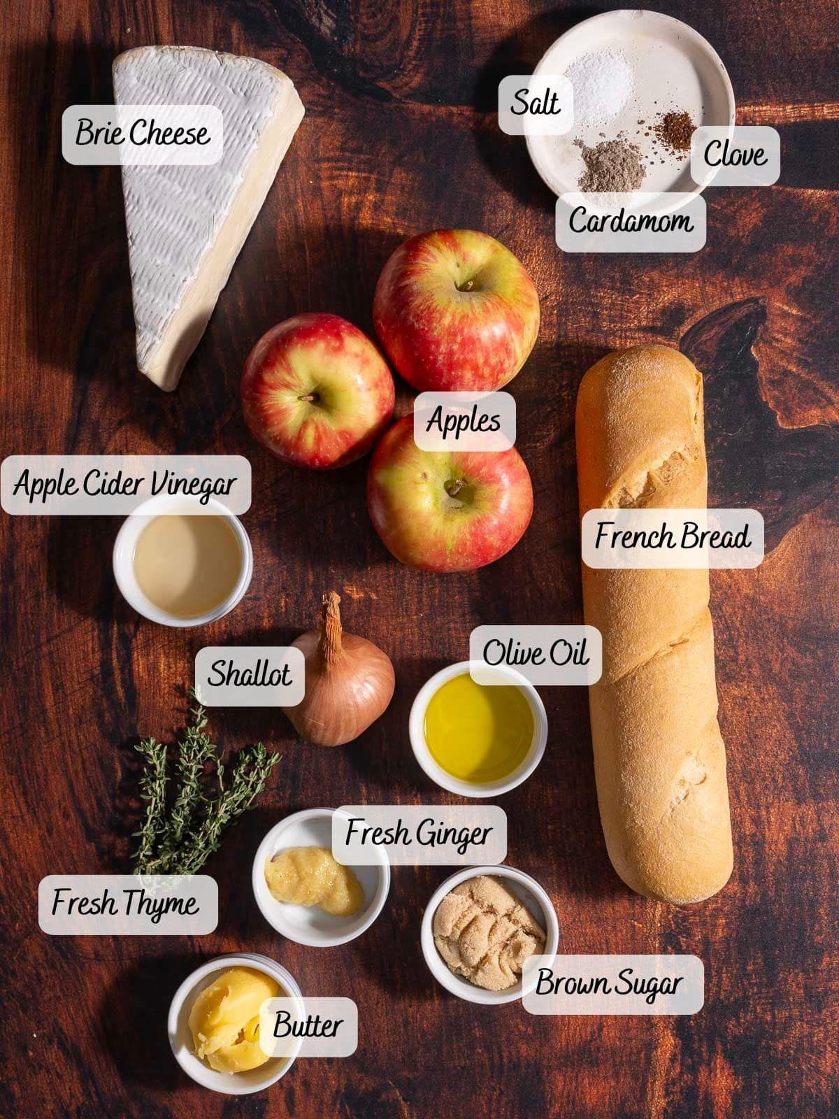 Ingredients are laid out on a wooden surface: ripe apples, a wedge of creamy Brie cheese, French bread perfect for crostini, apple cider vinegar, a shallot, olive oil, fresh ginger, brown sugar, butter, fresh thyme, salt, clove, and cardamom.