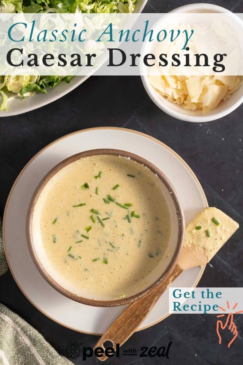 Bowl of Caesar dressing garnished with herbs, with a small dish of grated cheese and a plate of chopped greens in the background. Text: "Classic Anchovy Caesar Dressing.