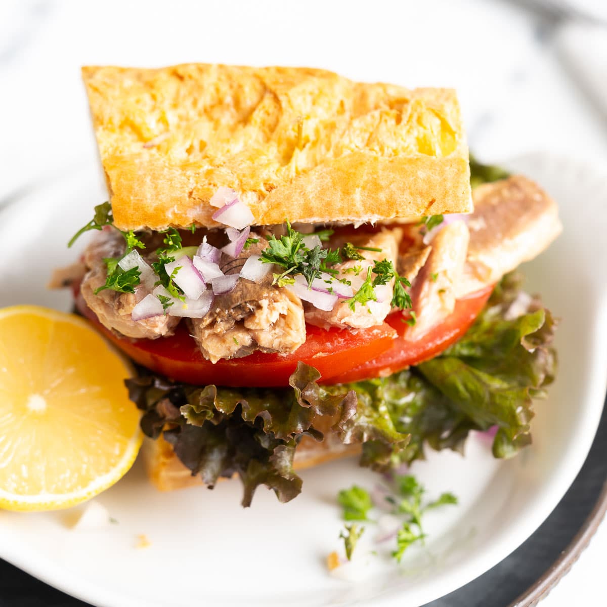 A sandwich with canned sardines, tomato slices, lettuce, and chopped onions on a square bread, garnished with parsley, is served on a plate with a lemon wedge.