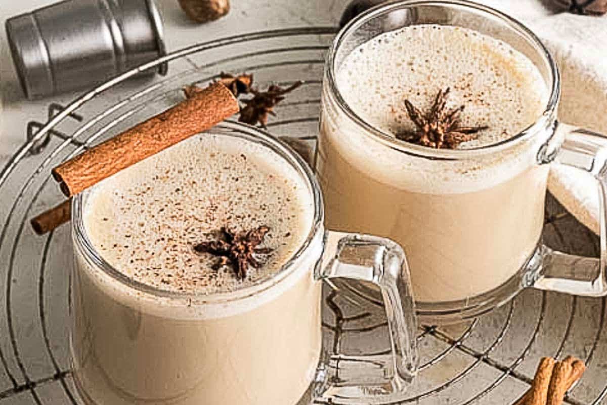 Two glass mugs of spiced eggnog, topped with star anise and cinnamon sticks, sit on a metal cooling rack-perfect cozy cocktails to serve as Thanksgiving drinks.