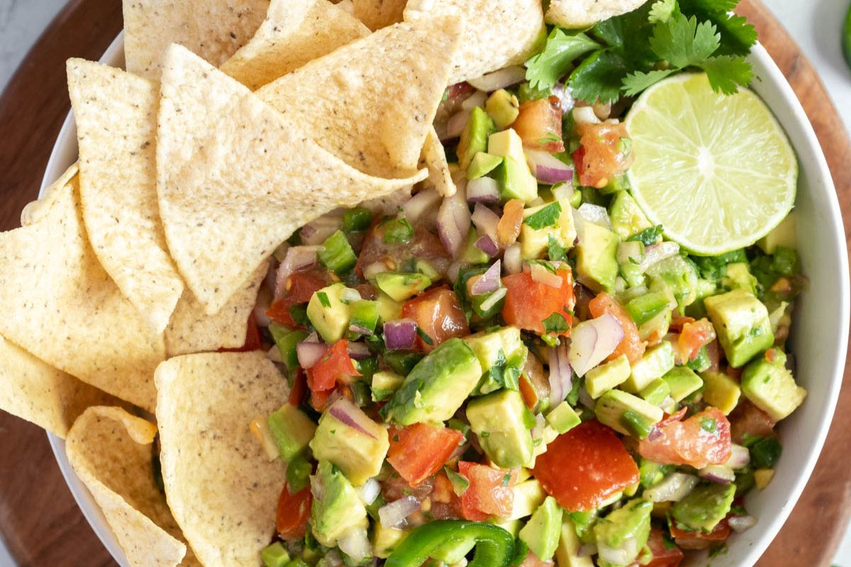 A bowl of fresh guacamole with diced avocado, tomatoes, onions, and cilantro, garnished with a lime wedge, next to tortilla chips.