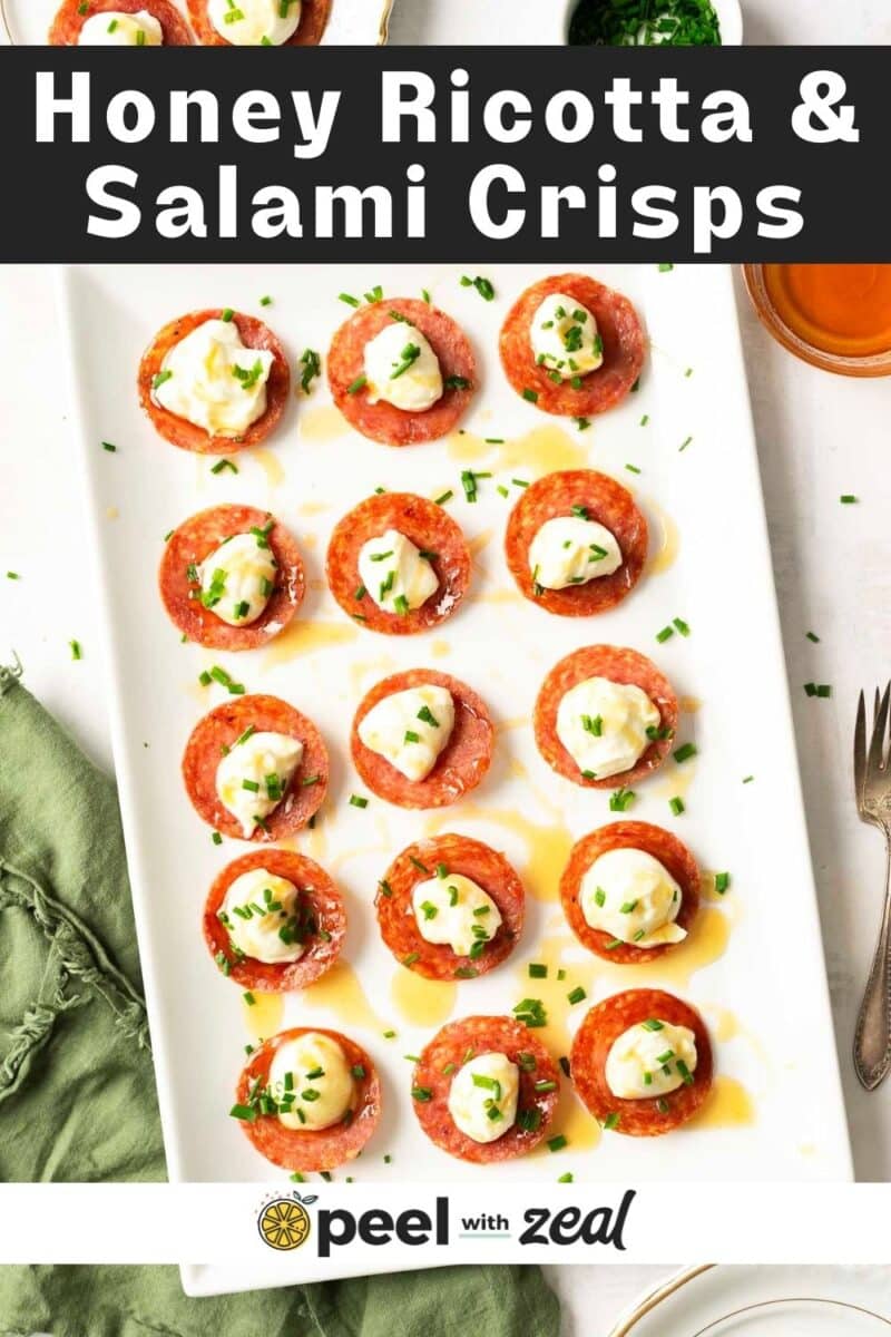 Plate of honey ricotta and salami crisps garnished with herbs, placed on a white rectangular dish. A green napkin and utensil are nearby.