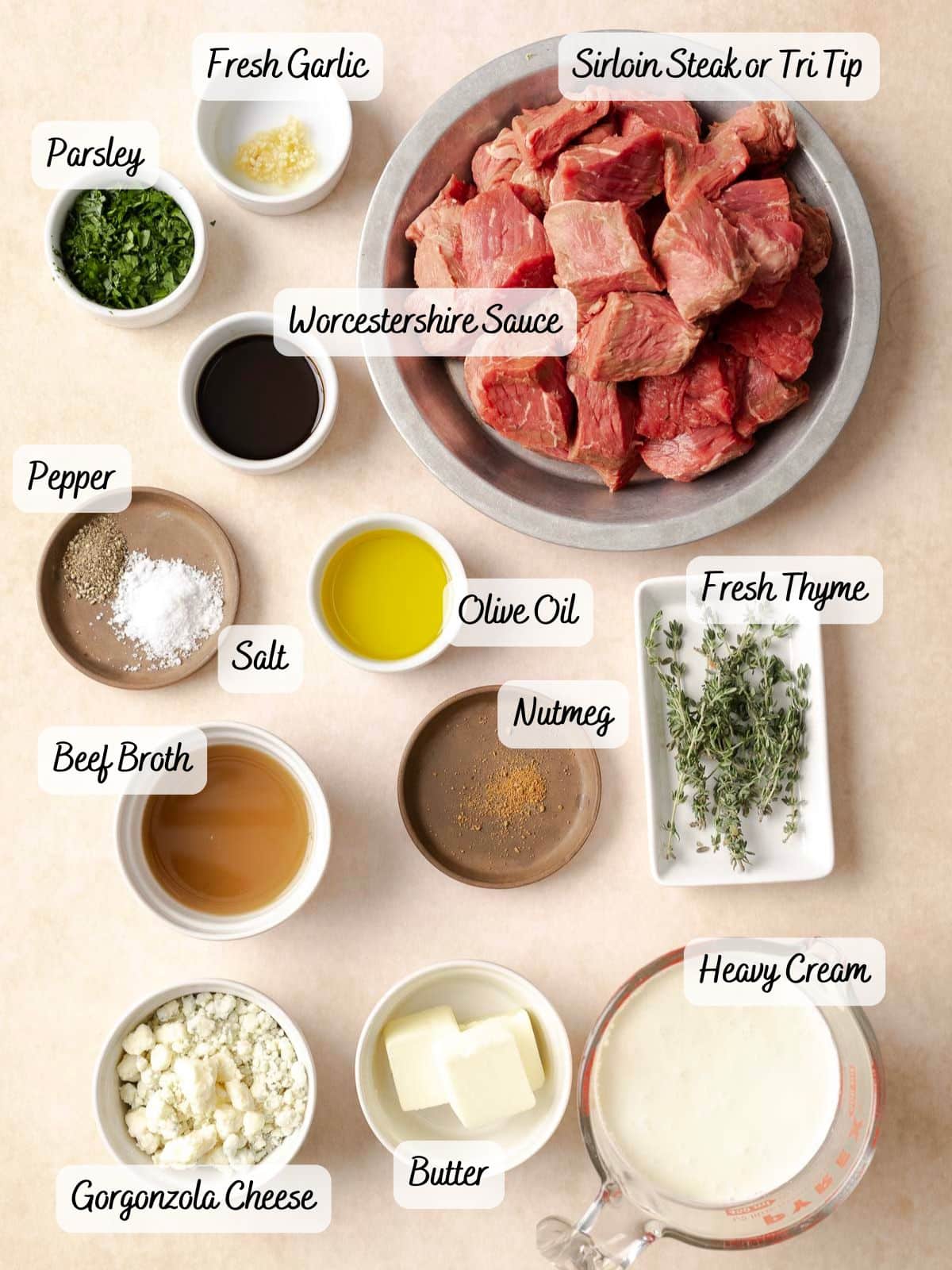 Overhead view of ingredients including steak, Worcestershire sauce, fresh garlic, parsley, pepper, olive oil, fresh thyme, nutmeg, heavy cream, beef broth, salt, gorgonzola cheese, and butter.