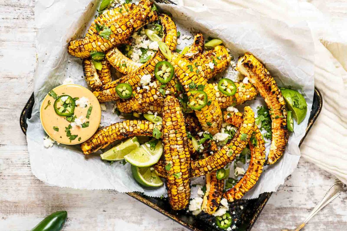 Grilled corn ribs topped with cheese, jalapeños, and cilantro, served with lime wedges and dipping sauce in a tray.