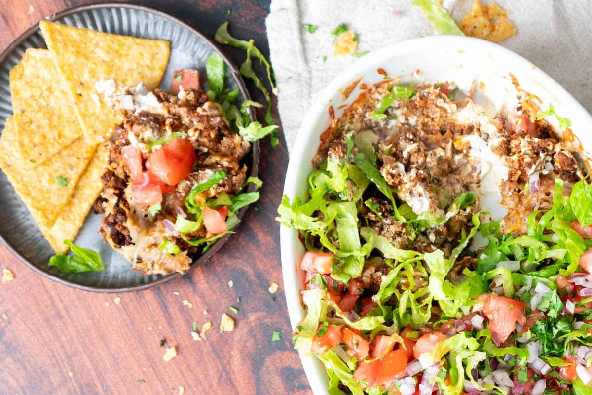 A plate of tortilla chips next to a dish filled with layered taco dip, topped with lettuce, tomatoes, and onions.