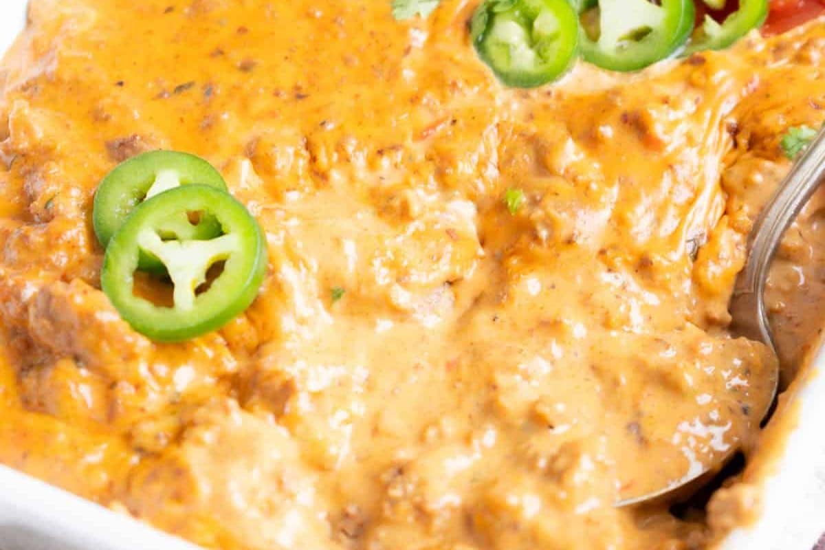 Close-up of creamy cheese dip with ground meat, topped with sliced jalapeños and a spoon in the corner.