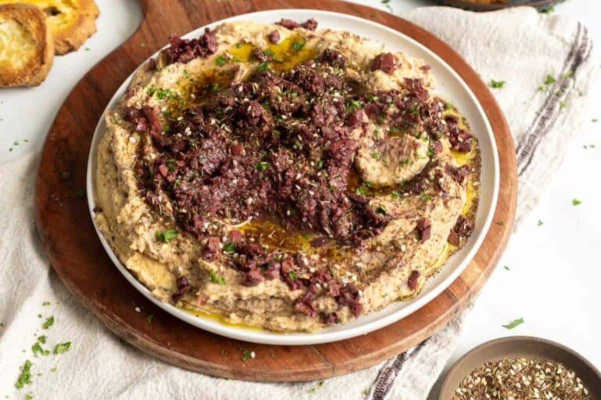 A plate of hummus topped with chopped kalamata olives and herbs, drizzled with olive oil, on a wooden board.