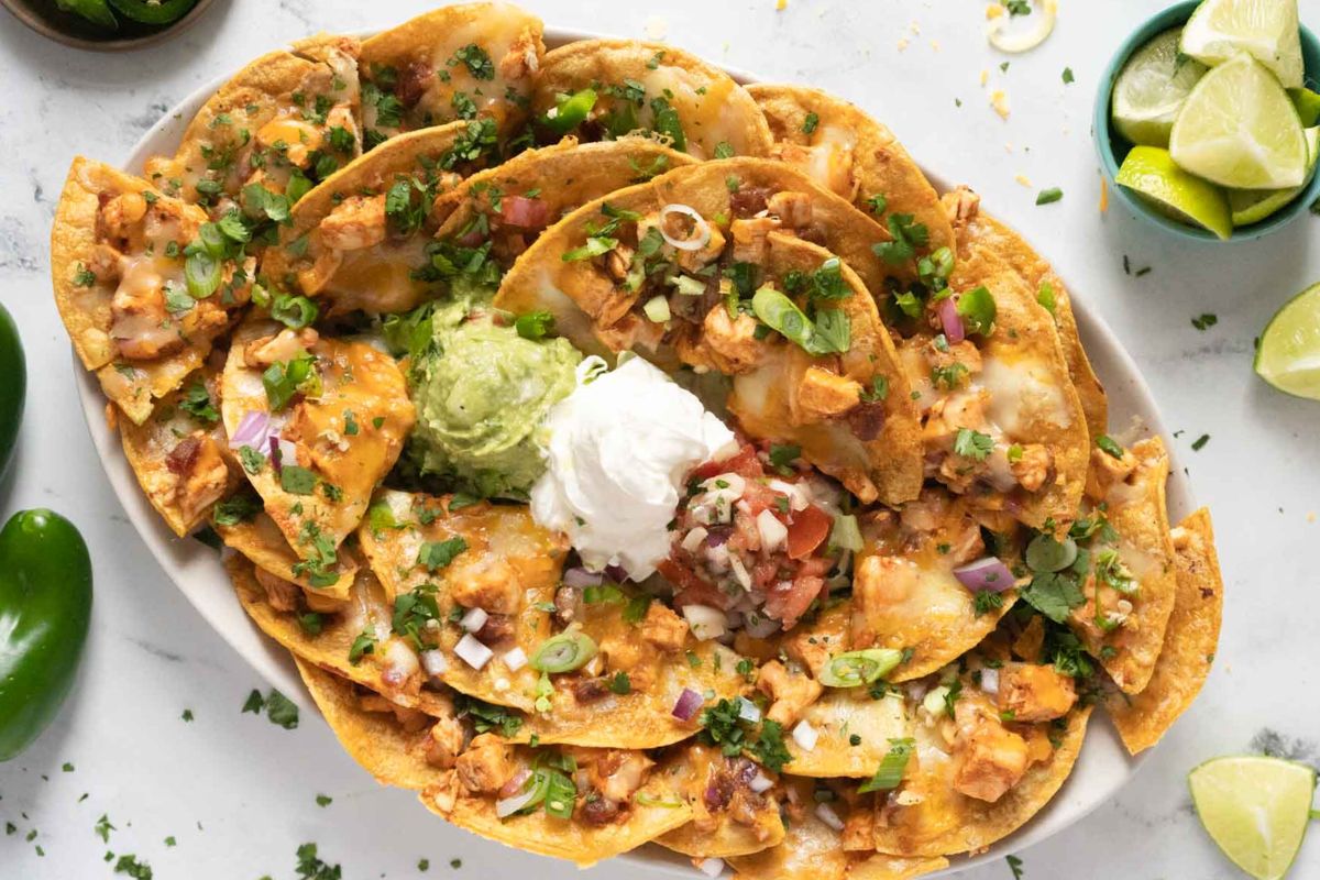 Plate of loaded nachos topped with cheese, chicken, green onions, tomatoes, guacamole, and sour cream. Lime wedges and jalapeño are on the side.
