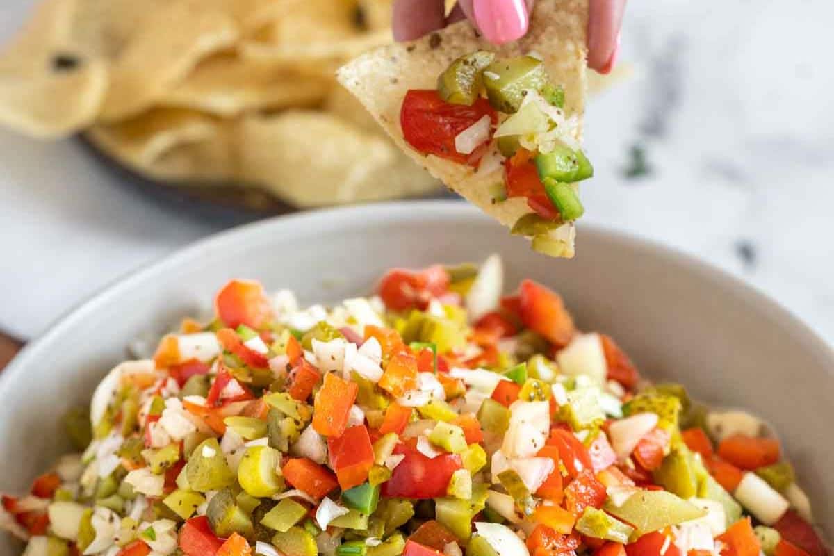 Person holding a tortilla chip with a colorful salsa made of diced vegetables, above a bowl with more salsa.