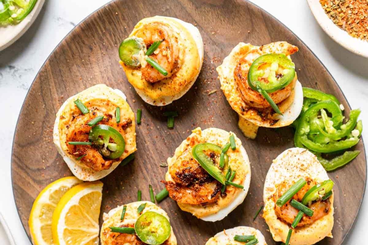 Deviled eggs topped with shrimp, jalapeño slices, and chives on a wooden plate, garnished with lemon slices.