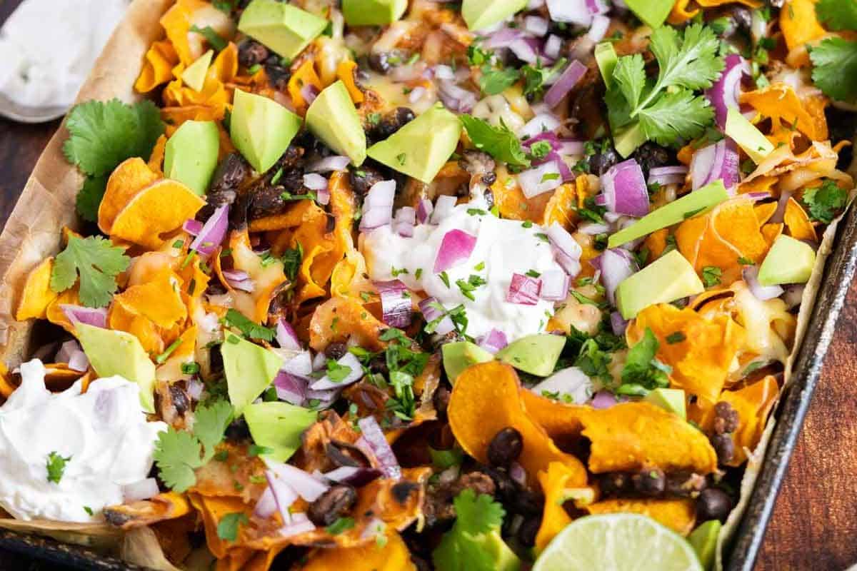 Colorful nachos topped with avocado, black beans, red onion, cheese, cilantro, and dollops of sour cream on a tray. Lime wedge on the side.