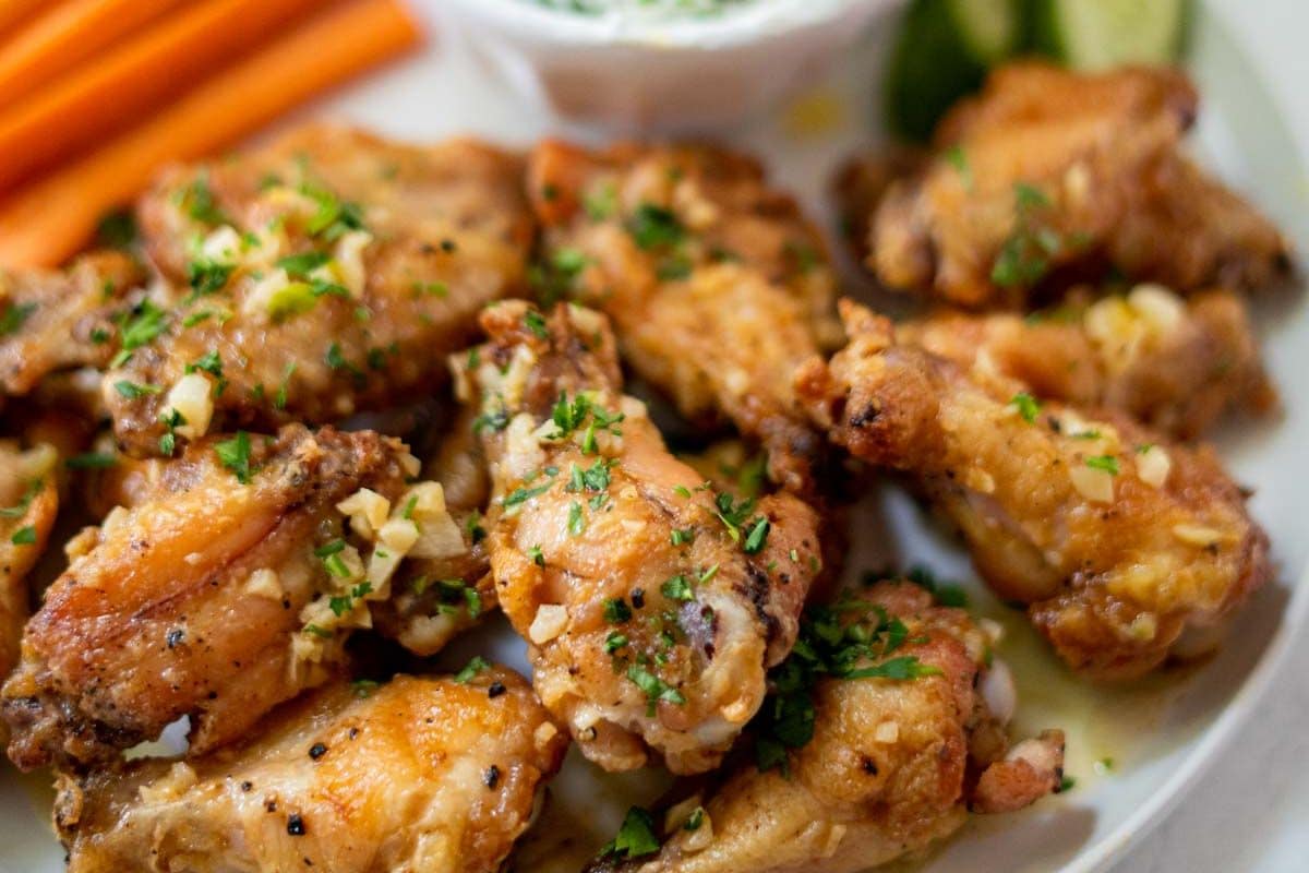 A plate of seasoned chicken wings garnished with herbs, accompanied by dipping sauce and sliced vegetables.