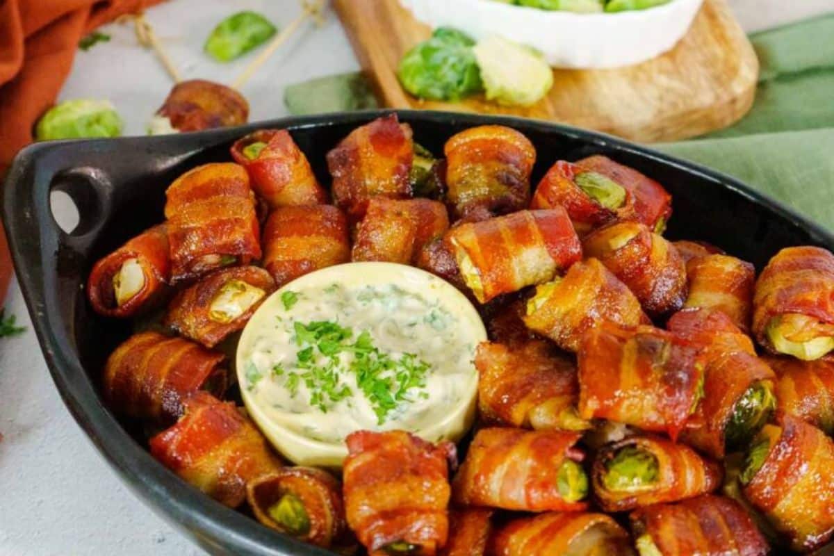 A black dish with bacon-wrapped Brussels sprouts surrounding a bowl of creamy dip, garnished with parsley.