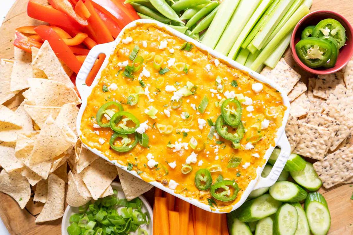 A colorful spread featuring a dish of cheesy dip topped with jalapeños, surrounded by tortilla chips, bell pepper strips, snap peas, celery, cucumber slices, crackers, and carrot sticks.