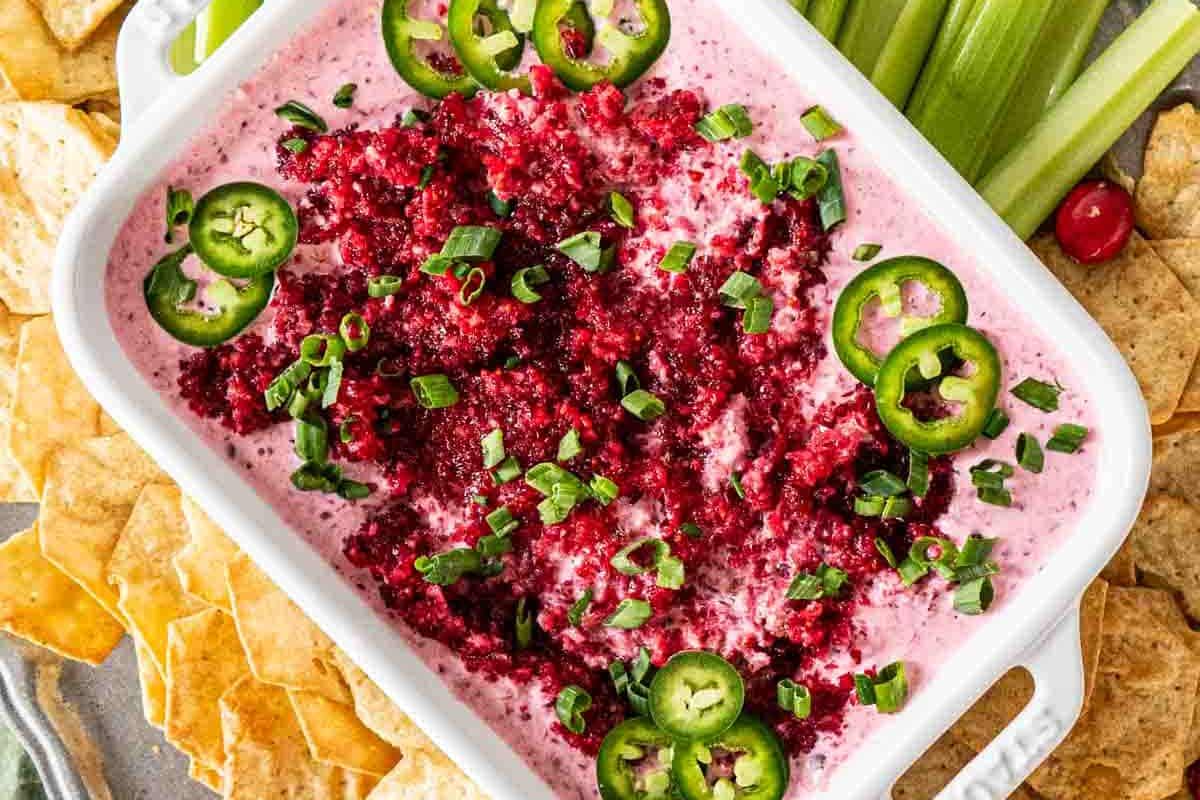 A dish with creamy pink dip topped with chopped green onions, jalapeño slices, and a red garnish, surrounded by celery sticks and pita chips.