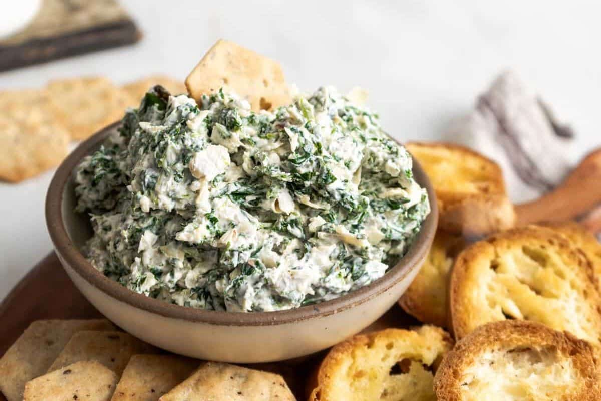 A bowl of spinach and artichoke dip surrounded by crackers and toasted bread slices on a wooden platter.