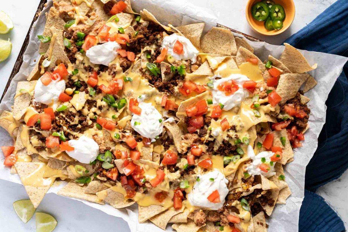 A tray of nachos topped with melted cheese, ground meat, diced tomatoes, green onions, jalapeños, and dollops of sour cream. Lime wedges and sliced jalapeños are on the side.
