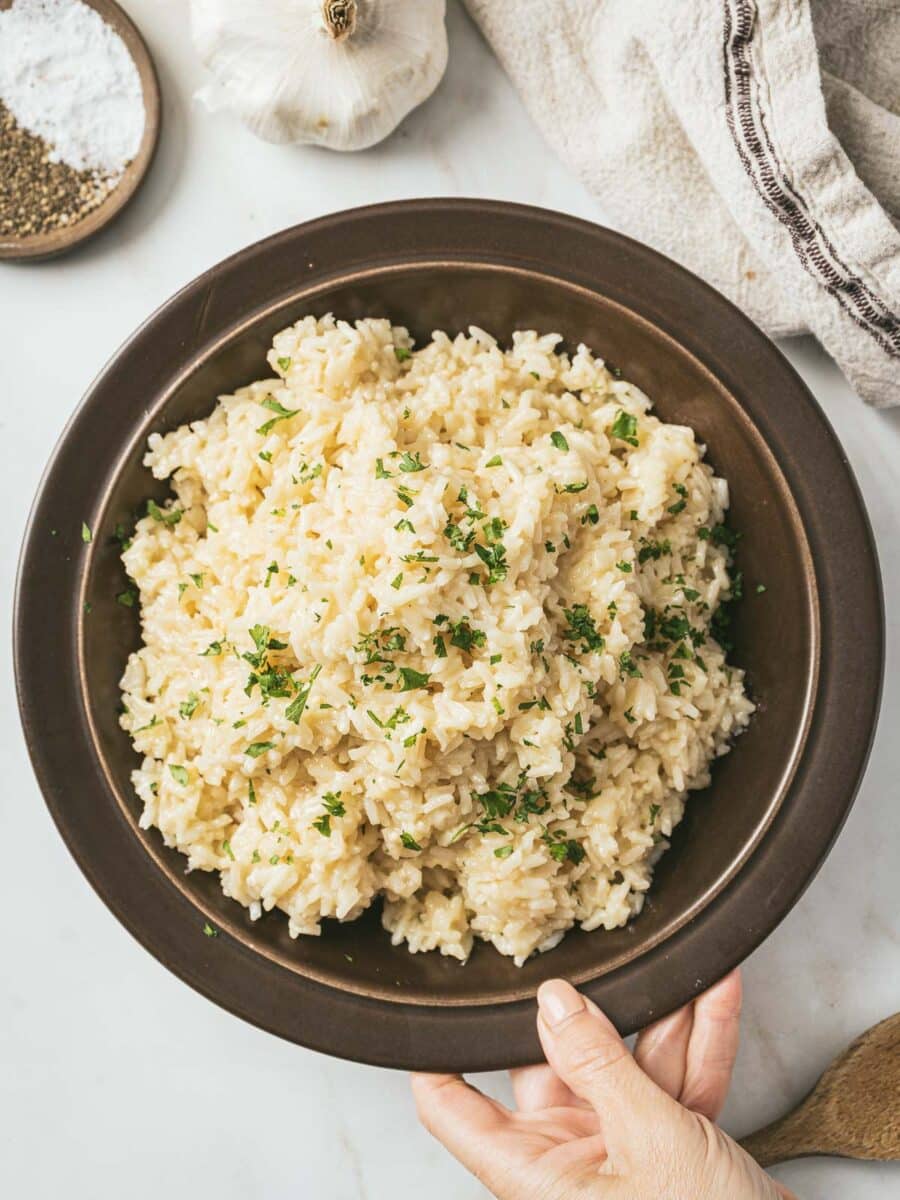 A hand holding a brown bowl filled with garlic parmesan rice garnished with herbs. A garlic bulb, towel, and spices are nearby on the white surface, making it a perfect addition to your array of rice side dishes.
