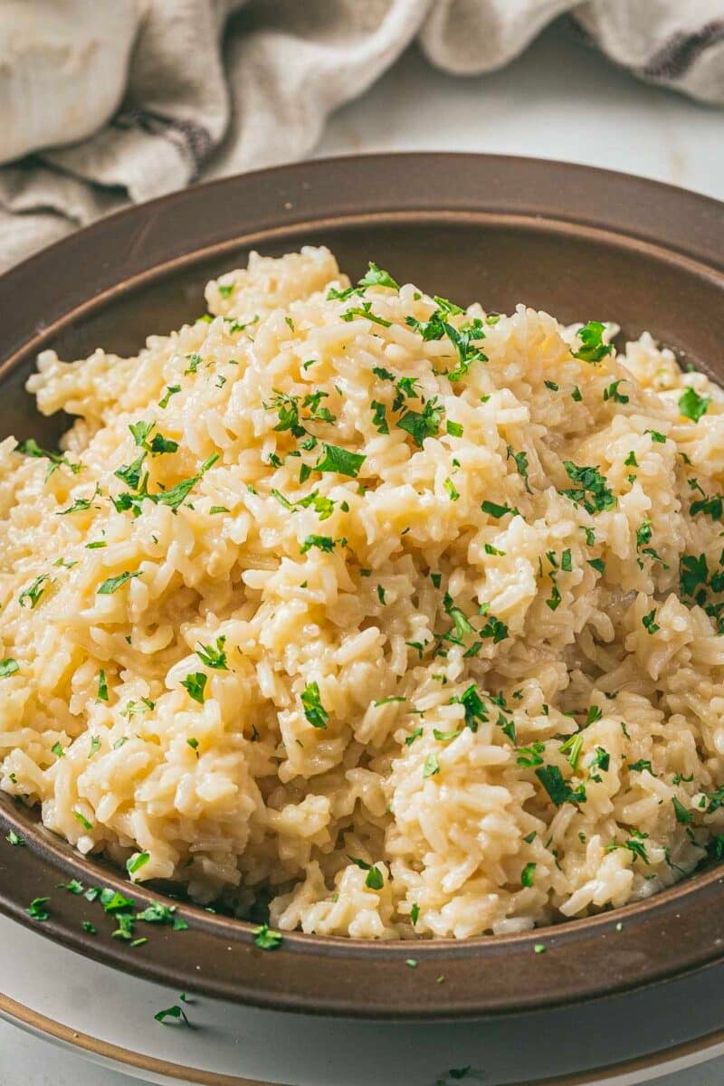 A bowl of creamy garlic parmesan risotto garnished with chopped parsley sits elegantly on a brown plate, making it a delightful rice side dish.