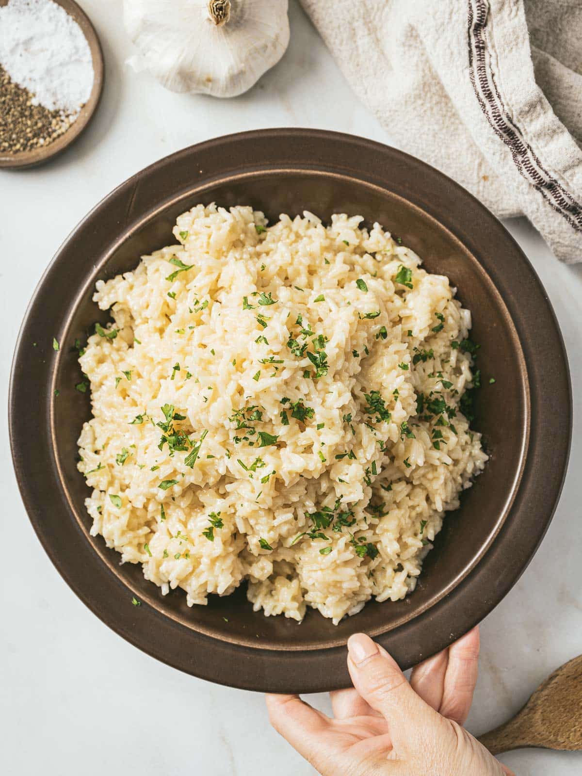 A hand holding a brown bowl filled with garlic parmesan rice garnished with herbs. A garlic bulb, towel, and spices are nearby on the white surface, making it a perfect addition to your array of rice side dishes.