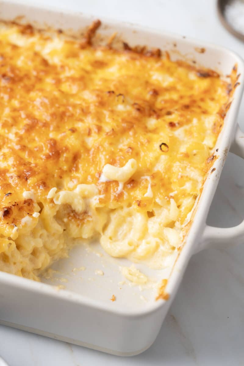 A baked macaroni and cheese dish in a rectangular white baking dish with a portion missing, revealing creamy pasta and a golden, crispy cheese topping.