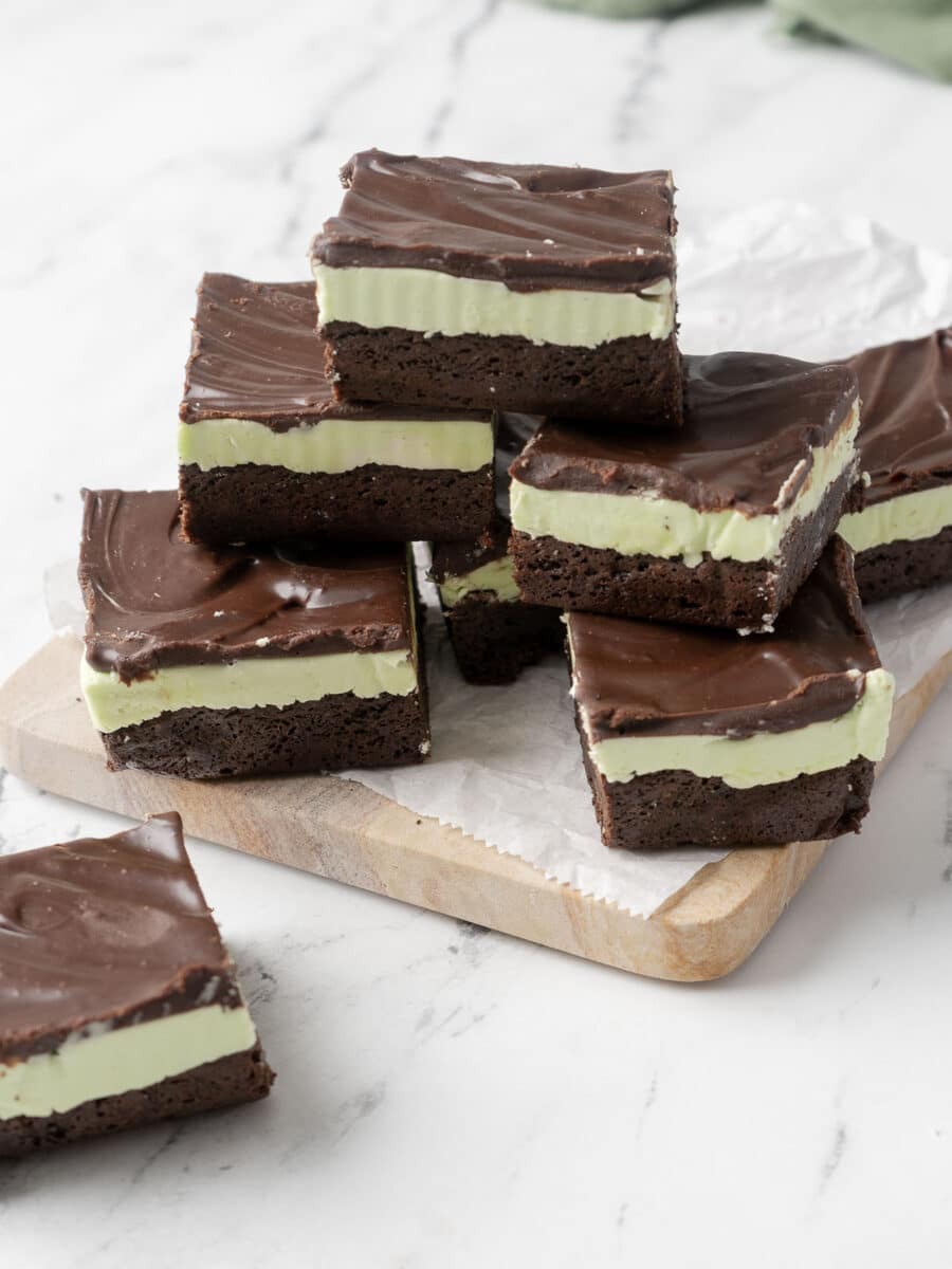 A wooden board showcases stacked mint chocolate brownies, featuring layers of rich chocolate cake, refreshing mint cream, and decadent chocolate topping. These delightful treats are gluten-free, ensuring that everyone can indulge in their irresistible charm.