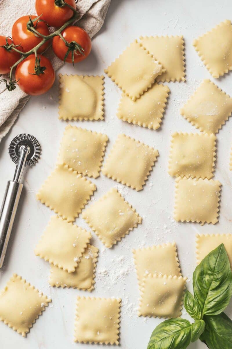 Fresh, gluten-free ravioli rests on a floured surface with a pasta wheel, vine tomatoes, and basil leaves nearby.