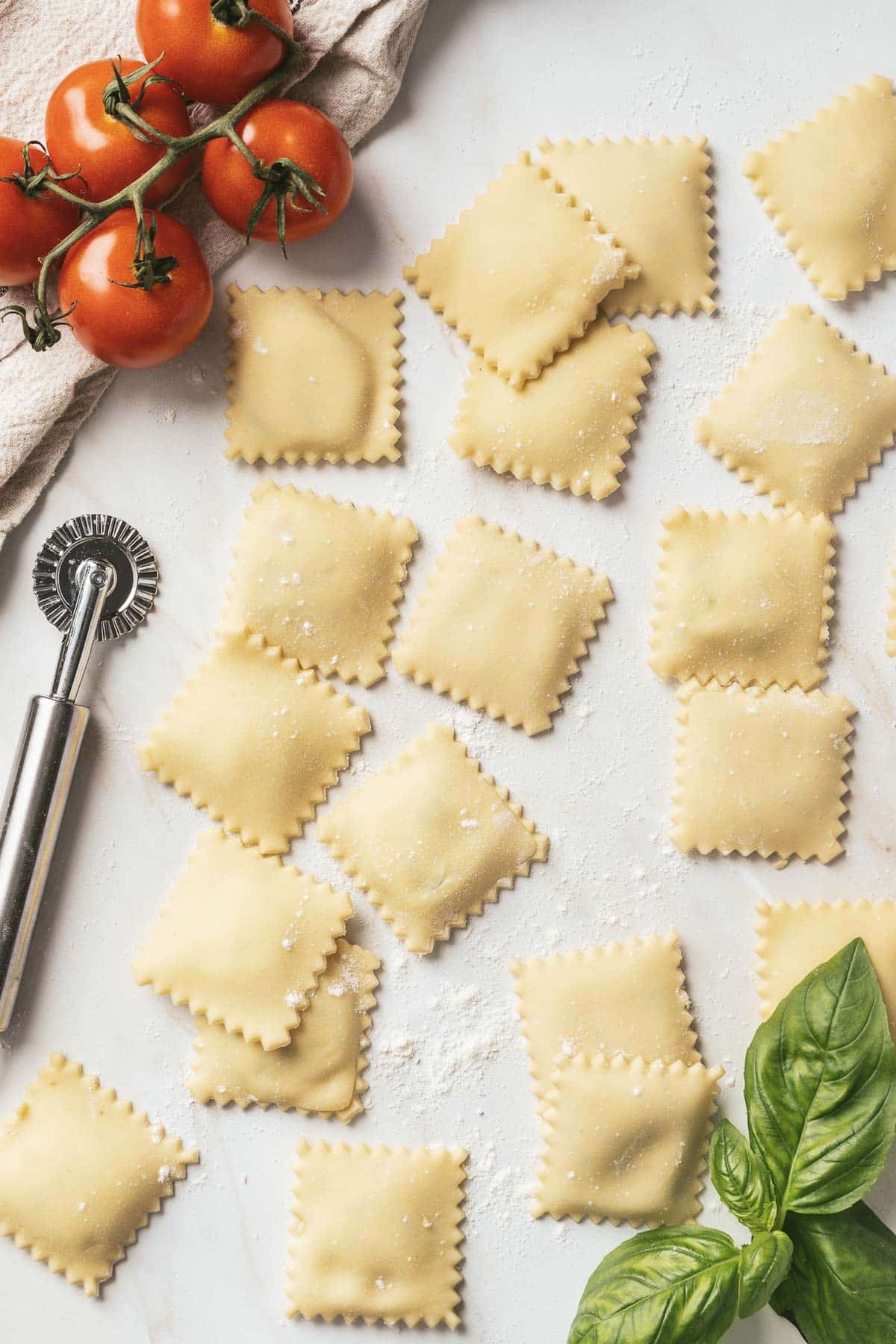 Fresh, gluten-free ravioli rests on a floured surface with a pasta wheel, vine tomatoes, and basil leaves nearby.