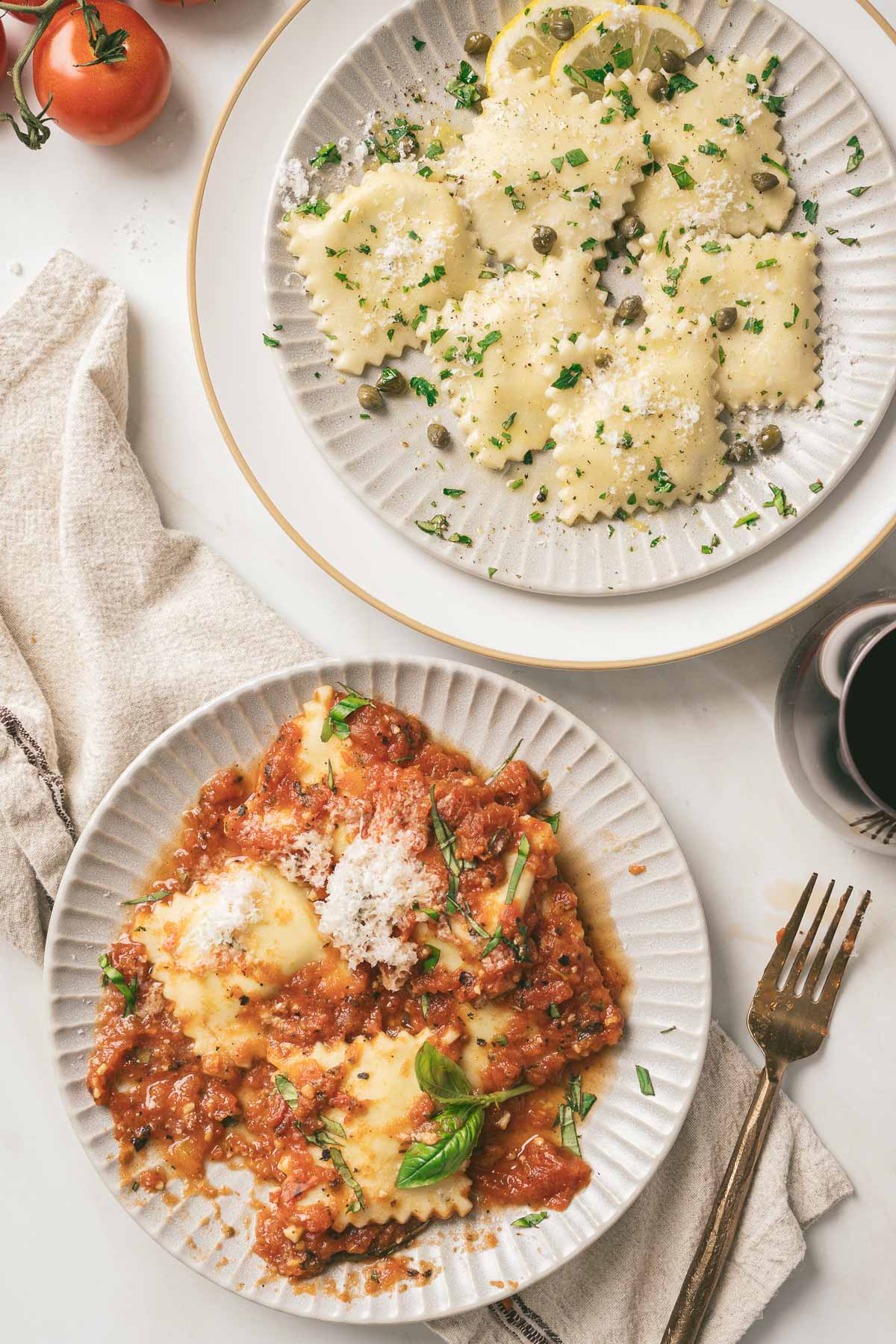Two plates of gluten-free ravioli: one with a creamy sauce, capers, and parsley, the other with tomato sauce, parmesan, and basil. A fork, linen napkin, tomatoes, and a glass are nearby.