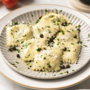 A plate of gluten-free ravioli garnished with capers, chopped herbs, and grated cheese, with lemon slices on the side.