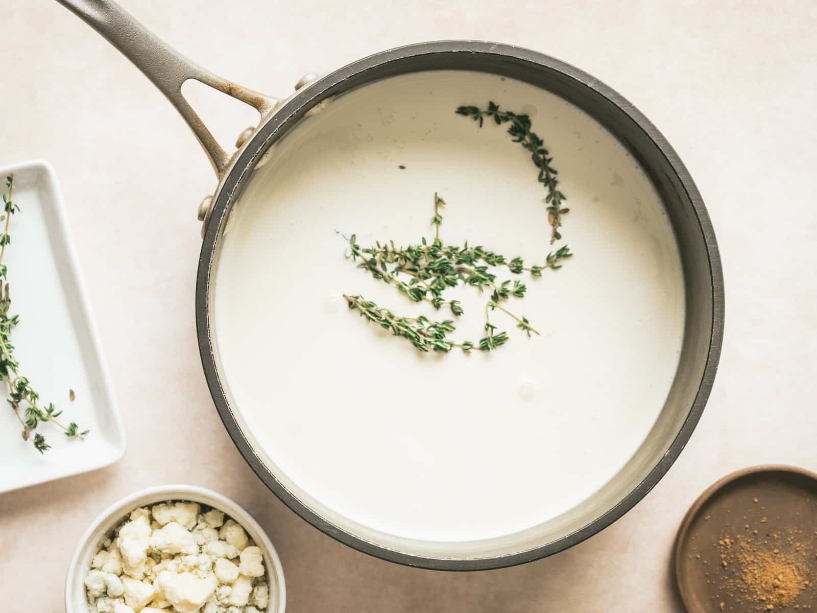 A saucepan filled with creamy white sauce, gently infused with Gorgonzola, is garnished with sprigs of thyme. Nearby are a dish of crumbled cheese and an empty brown dish lid.