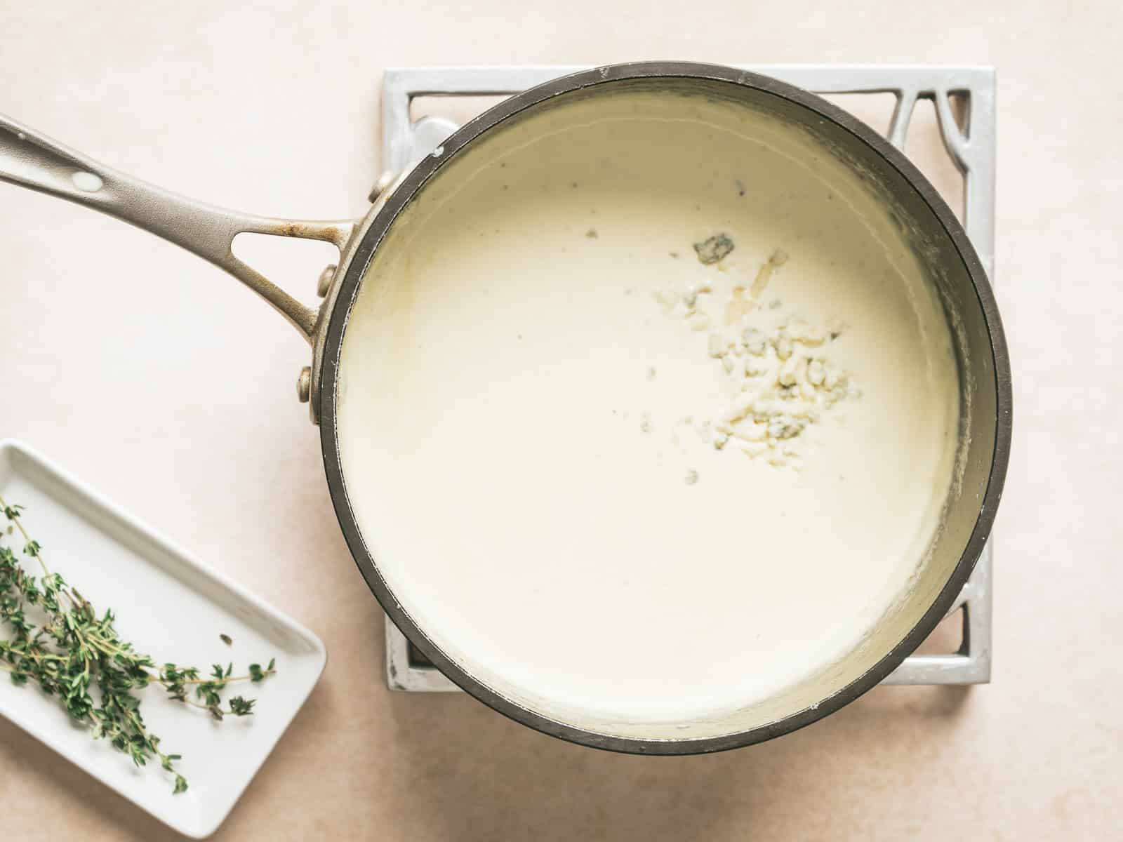 A saucepan with creamy Gorgonzola sauce simmers gently on the stove. A few herb sprigs are elegantly placed on a small rectangular dish beside it.