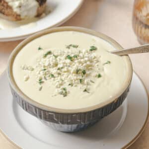 A bowl of creamy gorgonzola sauce topped with crumbled Gorgonzola and fresh herbs A plate with bread is partially visible in the background.