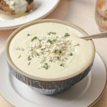 A bowl of creamy gorgonzola sauce topped with crumbled Gorgonzola and fresh herbs A plate with bread is partially visible in the background.