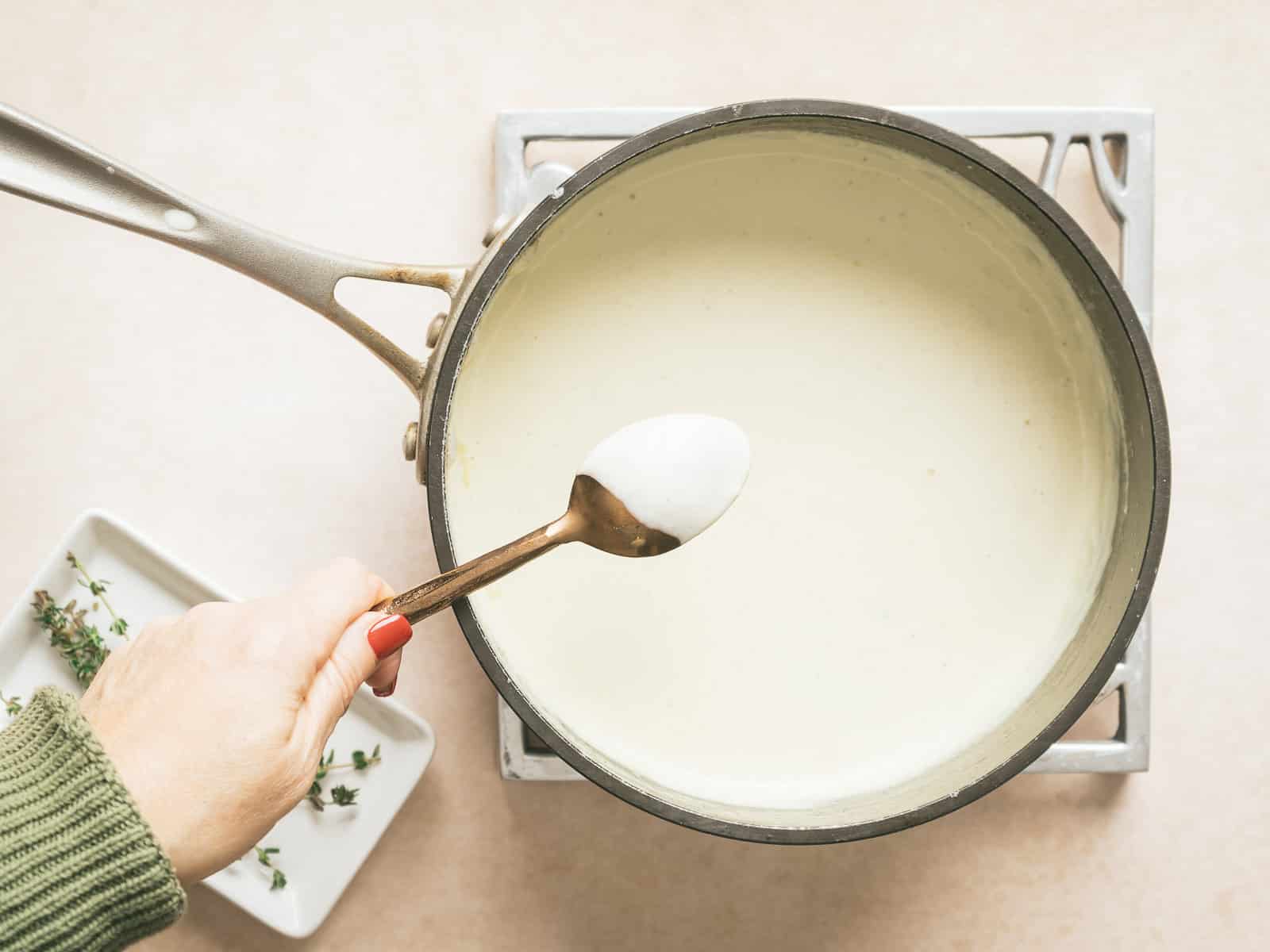 A hand holding a spoon hovers over a saucepan filled with creamy Gorgonzola sauce on the stovetop, accompanied by a small dish of herbs nearby.