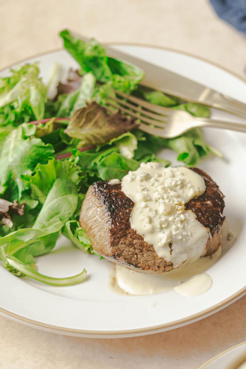 A grilled steak topped with a rich Gorgonzola cream sauce is served on a white plate next to a green salad with leafy vegetables. A fork and knife rest on the plate's edge.