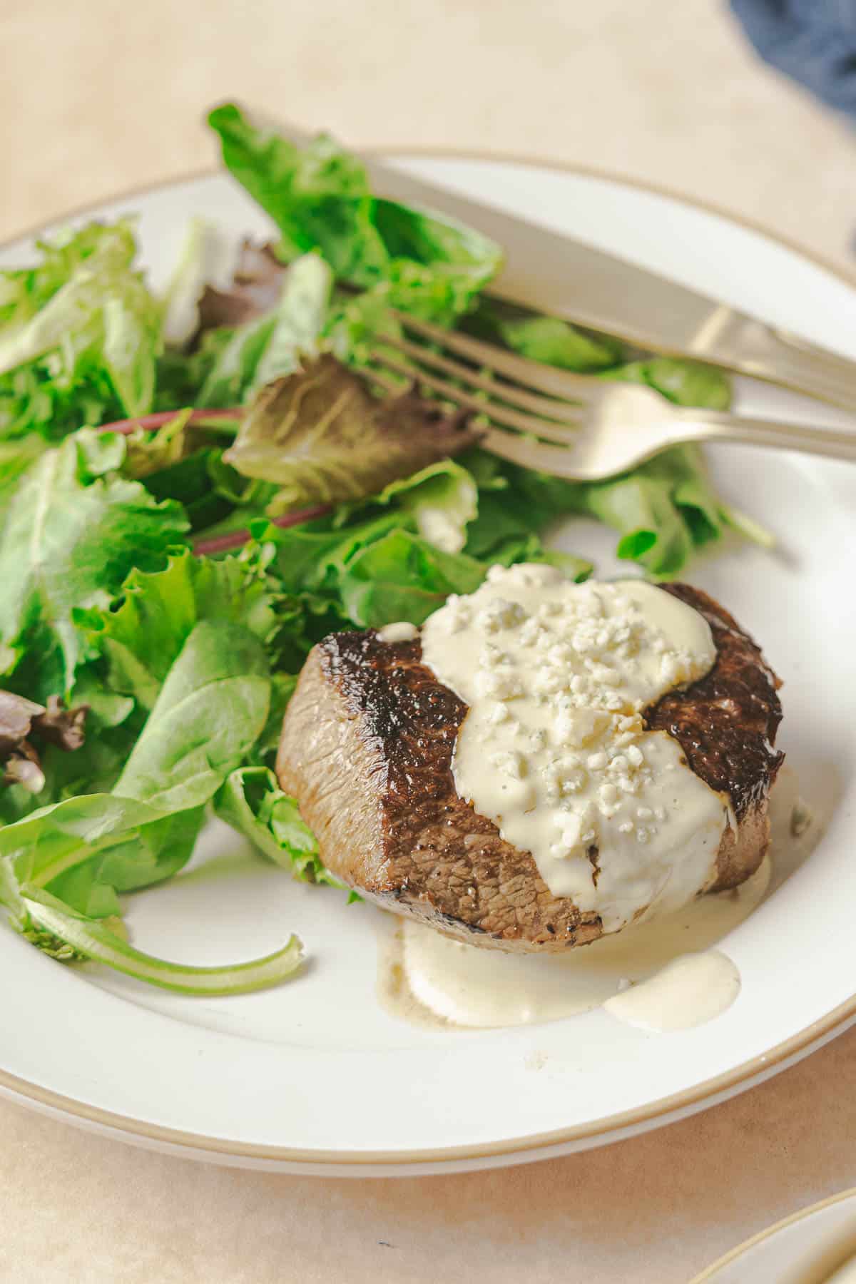 A grilled steak topped with a rich Gorgonzola cream sauce is served on a white plate next to a green salad with leafy vegetables. A fork and knife rest on the plate's edge.