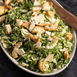 A Caesar salad featuring kale and shredded Brussels sprouts, topped with croutons and cheese shavings on a platter.