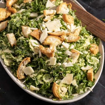 A Caesar salad featuring kale and shredded Brussels sprouts, topped with croutons and cheese shavings on a platter.